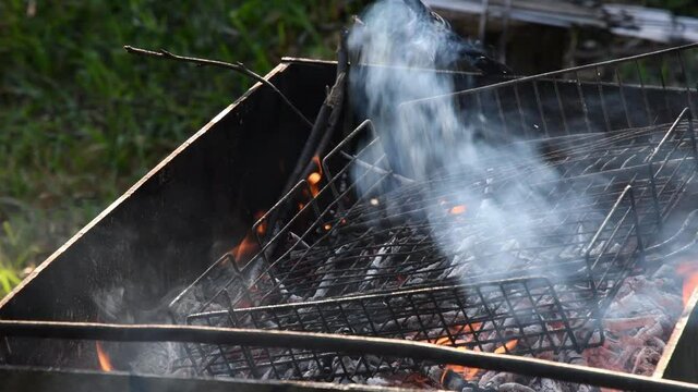 Fire burning in grill with old metal grid and messy smoldering coals and twigs covered with ashes and grey flying smoke. Flames and charcoals inside rustic mangal outdoors