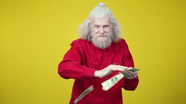 Portrait of wealthy senior man scattering money looking at camera. Rich Caucasian old retiree with long grey hair posing with cash at yellow background. Richness and wealth concept.