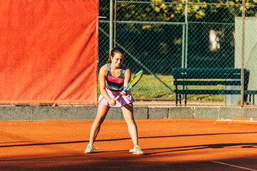 A young fit woman plays tennis outdoor on an orange tennis field early in the morning