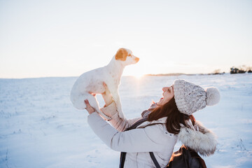 happy woman in snowy mountain wearing modern coat at sunset. Holding cute jack russell dog in arms. winter season. nature at sunset