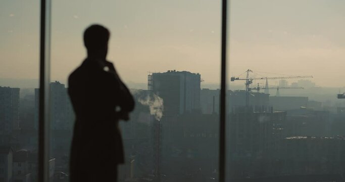 Silhouette of successful businessman contemplating business idea looking through large window at city buildings. Modern cityscape. Sunsets.
