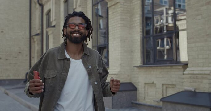 Joyful guy with dreadlocks smiling and dancing while looking to camera outdoors. Bearded afro american man in stylish red glasses having good mood and singing while walking at street