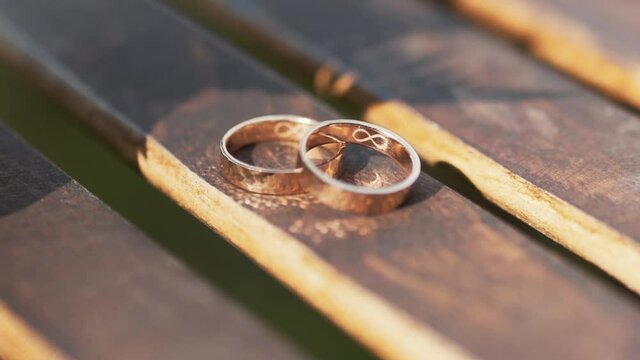 Gold wedding rings on inside of engraving of infinity symbol lie on a wooden rail in the sun rays