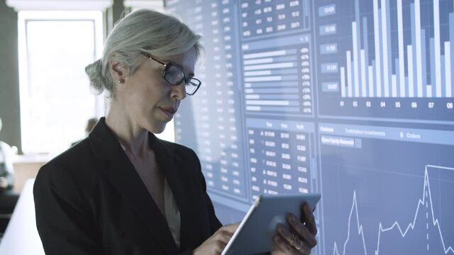 Middle-aged content businesswoman using tablet and standing near big screen in office. Professional successful manager preparing for presentation and checking charts. Business and management concept
