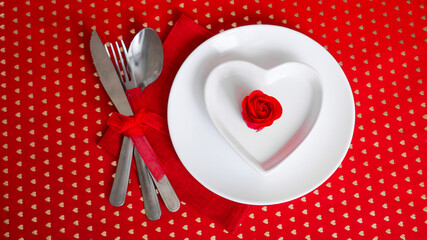 Romantic decoration of holiday table. White plates in the shape of a heart with a red rose. Red background. Top view