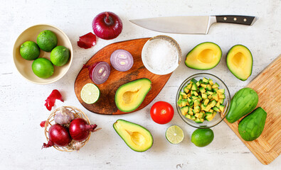 Avocado halves, pieces, tomato, limes and onions, cup with salt - basic guacamole ingredients and chef knife on white working board, flat lay photo