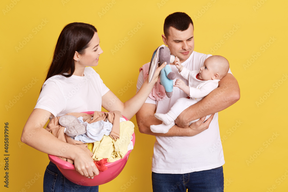 Wall mural Lovely young family of three being photographed against yellow wall, mommy doing landry and holding basin full of dirty clothing, father with infant in hands trying to comfort baby, mommy shows toy.