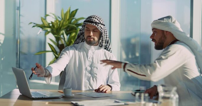 Two Handsome Successful Arabian Investors in Muslim Traditional Clothes Sitting in Office Using Laptop Computer Discussing Business Plans.