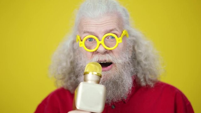 Headshot of positive bearded male retiree in funny eyeglasses singing at microphone on yellow background. Close-up portrait of cheerful carefree Caucasian senior man having fun enjoying karaoke.