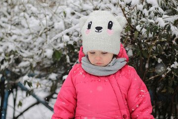 Little sad child in a red warm jacket and hat against the background of a snowy green bush