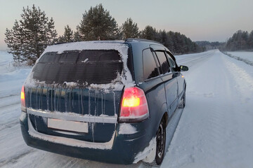 a frozen car stands on the side of a slippery winter road at dusk. Orange warning lights are on. The rear window was covered with ice, snow and icicles. Dangerous long journeys in the cold.