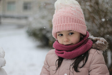 Lovely dark-haired little girl schoolgirl in winter clothes stands against the background of snow-covered blurry bushes