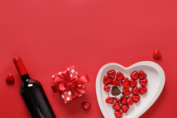 Valentine's day composition, gift, red wine, chocolate sweets and decoration on red background. Romantic invitation with copy space.