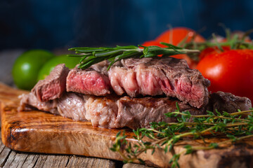 Medium rare grilled beef steak ribeye with rosemary and tomatoes on wooden cutting board on dark blue background. Appetizing image for restaurant menu. Delicious food. Juicy meat.