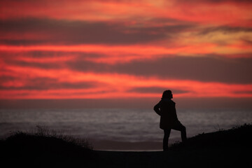 Fototapeta premium Silhouette of woman standing on hill overlooking the ocean with a dramatic orange sunset 