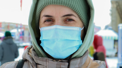 Fototapeta na wymiar Portrait of happy young woman wearing medical protective mask outdoors and looking at camera. Virus protection. A woman outdoors on a winter day stands in a hat and a hood in the park.
