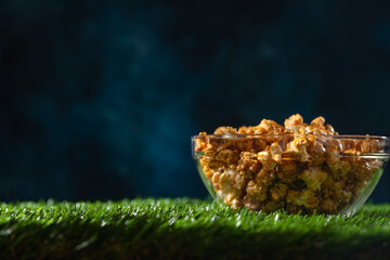 Glass bowl with caramel popcorn stands on the grass on dark blue background. Movie concept. Open-air cinema.