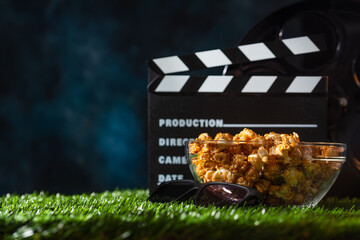 Movie clap board and glass bowl with caramel popcorn stands on the grass on dark blue background. Movie concept. Open-air cinema.