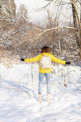 Back view of a woman in a bright yellow jacket and jeans with a backpack in a snowy landscape forest walks through the snowdrifts