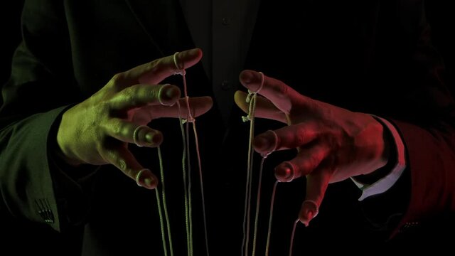 Businessman leader in a suit manipulates the strings of puppets tied to his fingers. Domination, power and submission concept. Close up mans hands illuminated with colored lights. Slow motion.