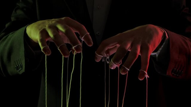 Puppeteer manipulating and pulling strings of marionettes. Male hands close up painted with colored lights on black background. People are toys in hands of dominant businessman dictator. Slow motion.