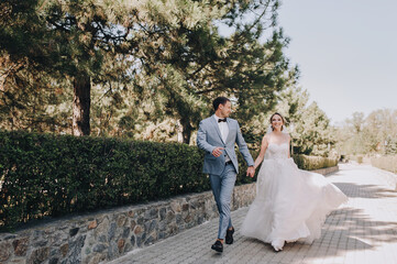 The groom in a gray checkered suit and the blonde bride in a white lace dress run holding hands...