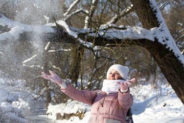 Woman in winter clothes on a walk in the park. Throws up a handful of snow. A river flows nearby. There is a lot of snow around.