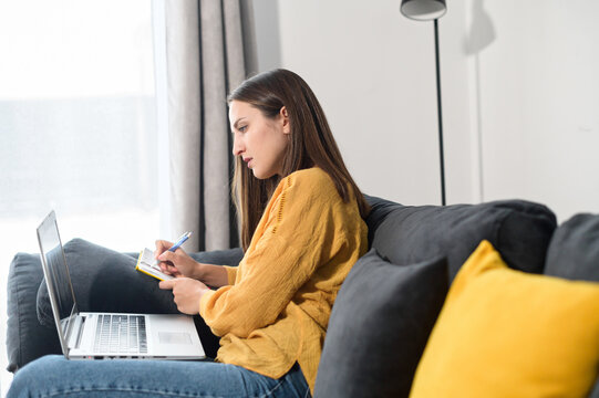 Focused young woman works with a laptop sitting on the couch at home, female student watching a webinar, video courses and taking a notes in a notepad. A female entrepreneur writing plans with a pen
