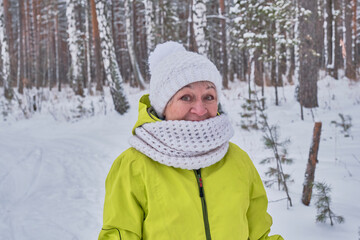 Senior woman in a yellow jacket, white knitted hat and scarf looking at the camera in surprise over the background of a winter snowy forest