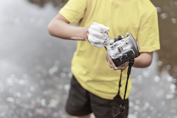 A boy in a yellow t-shirt and shorts is standing near the water in nature and washing a camera with...