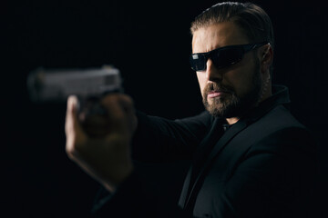 Bearded killer in sunglasses and suit standing over black studio background with real gun in hands. Aggressive bandit with weapon.