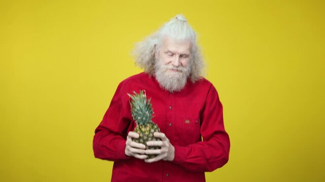 Joyful senior man dancing with pineapple at yellow background. Portrait of cheerful old Caucasian guy having fun. Leisure and joy concept.