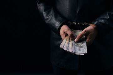 Close up of male hands in handcuffs holding money cash. Arrested person with dollars over black...