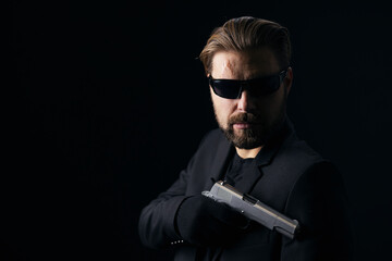Portrait of dangerous killer in sunglasses and gloves posing with real gun over black background. Bearded man aiming with weapon in dark frightening atmosphere.