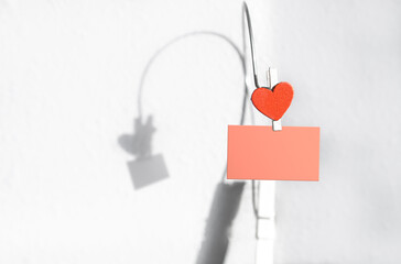 white wooden clothespin with red heart shape clip on blank pink paper note with blurred shadow on white wall background. Space of feeling expression. Valentine's day, concept of love and romantic