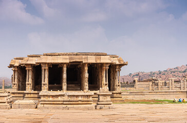 Obraz premium Hampi, Karnataka, India - November 5, 2013: Vijaya Vitthala Temple. Abandoned and ruinous mandapam hall on side with mountains on horizon under blue cloudscape. Female gardeners add color.