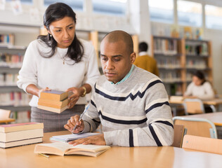 Female librarian helping focused adult hispanic student preparing to exam, bringing him books