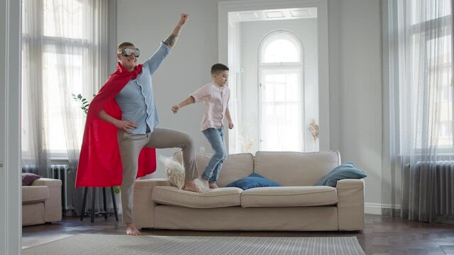 Father And Son In The Apartment Playing Game. Dad In Red Cloak And Glasses Depicts Superman.