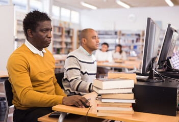 Focused young adult man sitting at table with computer and books, finding information at library