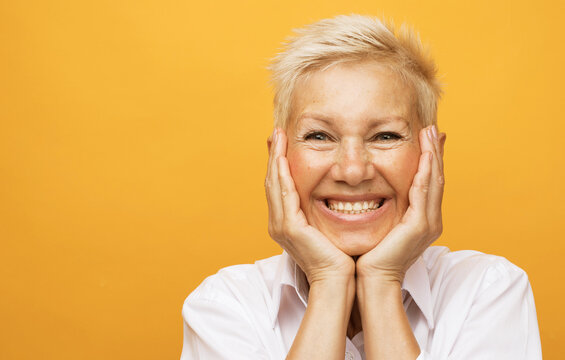 Life only starts when get older. Portrait of charming happy and carefree european senior woman with blond short hair laughing and amusement holding palms on cheeks over yellow background