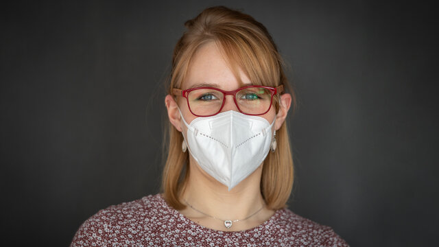 Rotglonde junge Frau mit Brille tr&auml;gt eine FFP2 Maske gegen das Coronavirus bzw. zum Schutz vor Covid 19- Panoramaformat isoliert vor dunkelgrauem Hintergrund mit viel Textfreiraum
