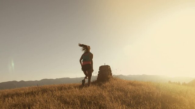 Slow motion shot of fit athlete woman running uphill and climbing small rocky moutain, picking view and throw her hands up in the air. Success.