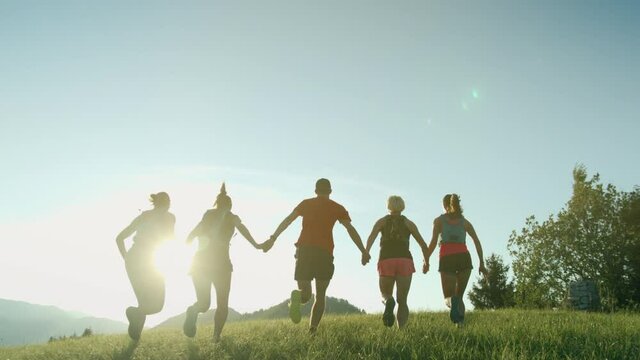 A team of runners running on top of the hill with mountain view in a beautiful golden sunset light. Runners holding their hands and together reach the goal. 