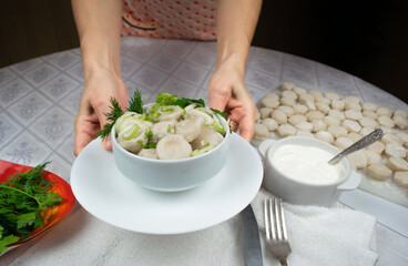 Hands of a woman in an apron hold out a bowl of cooked homemade dumplings, decorated with green leaves of dill and parsley. Selective focus.