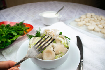 Bitten meat dumpling is pinned on a fork against background of bowl of cooked homemade dumplings with green dill and parsley leaves, sour cream and herbs. Close-up.