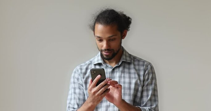 Smiling african bearded man in plaid shirt standing alone on gray studio background with smart phone wireless modern device reading media news, choosing goods purchasing on e-commerce web services