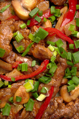 Fried meat with vegetables, green onions, bell peppers and mushrooms, in a frying pan, on a wooden background