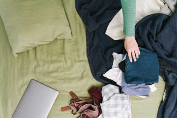 Woman reaching for laundry on messy bed with laptop