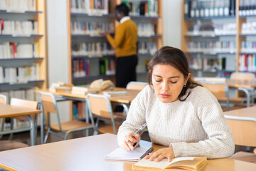 Focused young hispanic woman engaged in research, looking for information in books in public library