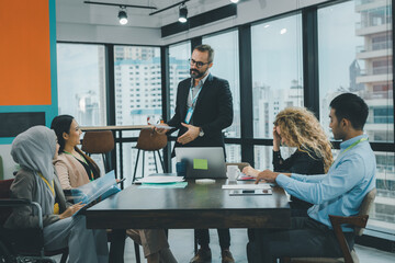 Chief executive male officer is standing at the head of the desk to introduce the new project. Group of employee are sitting and listening from their chief.